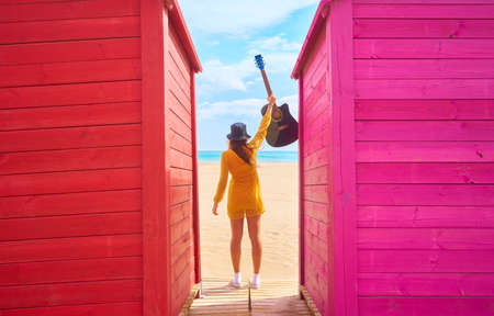 A backshot of a Caucasian teenager standing between colorful beach sheds and posing with an acoustic guitar - the concept of fun and leisureの写真素材