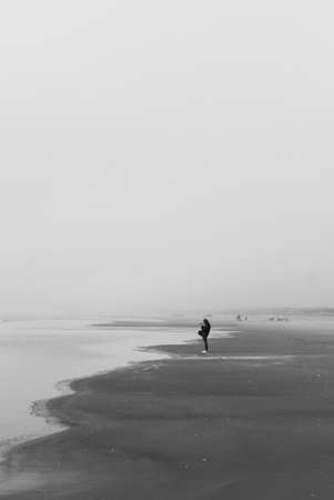 A grayscale shot of a lonely person walking on the beach under dark cloudsの写真素材