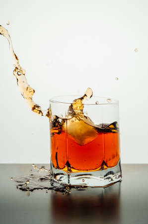 A vertical shot of a lowball glass of splashing whiskey with an ice piece on a reflective surface...