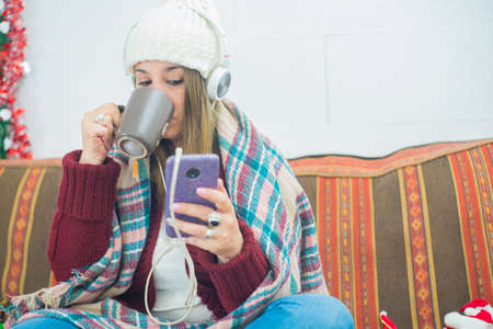 A closeup shot of a girl with headphones covered with a scarf drinking from a mug and looking at phone on couchの写真素材