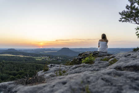 A back view shot of a young female sitting on the edge of a cliff and enjoying a majestic sunset over mountainsの写真素材