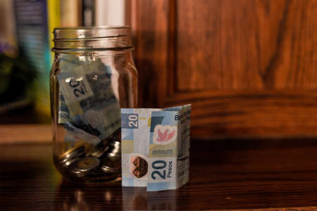 A closeup of coins and pesos in a jar on a wooden table under the lights with a blurry background