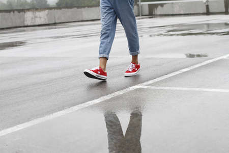 The legs of a person in jeans and red sneakers walking through the road after the rain