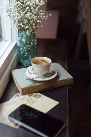 A cup of coffee on a book, a flower vase and a phone on a windowsillの写真素材