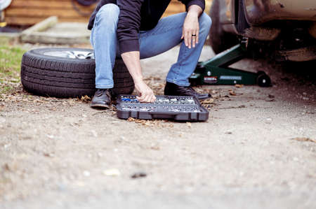 A shot of a man sitting on tire of  car and choosing a tool from a set