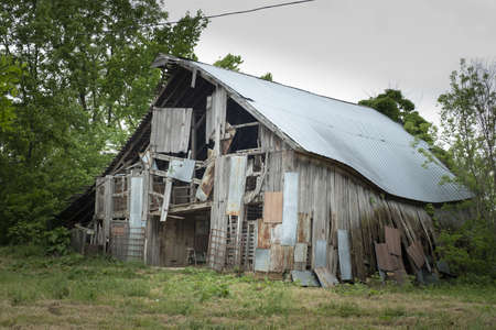 An eye-level shot of an old wooden decrepit barn standing near green treesの写真素材