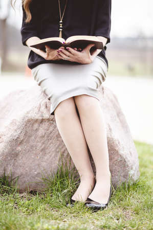 A vertical shot of a female wearing a grey skirt and reading a book while sitting on a rockの写真素材
