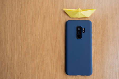A top view of a blue case smartphone with yellow paper boat above on a wooden table