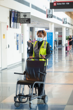 PHILADELPHIA, UNITED STATES - Jul 23, 2020: Airport Worker Rolling a Wheelchair Down a Philadelphia International Airport Concourse Wearing a Face Mask for COVID-19 Prevention.のeditorial素材