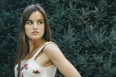 An attractive girl with brown long hair posing in front of the green trees during daytime