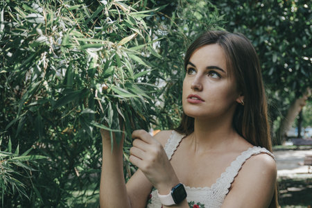 An attractive girl with brown long hair touching and looking up at the leaves of a green treeの写真素材
