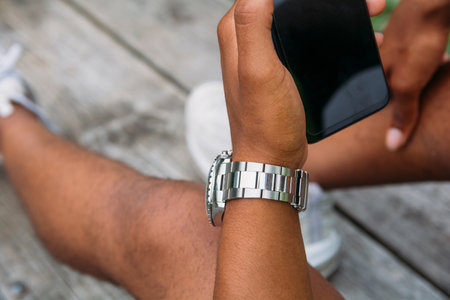 A young man with a watch on his wrist staring at his phone while sitting in a park