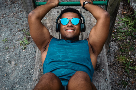 The young fit man with sunglasses and a blue shirt working out in the park while listening to music