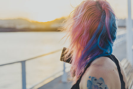 A young female with pink and blue hair and tattoos holding a camera standing near the seaの写真素材