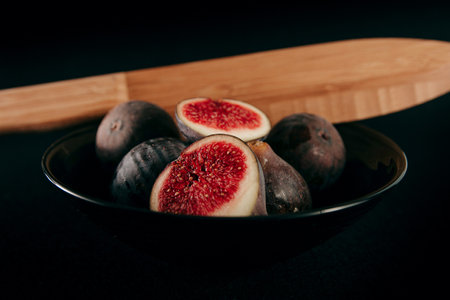A closeup shot of a few fresh figs in a black bowl with a wooden knife on a black backgroundの写真素材