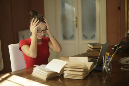 A young student with glasses studying in a room with books and laptops on a deskの写真素材