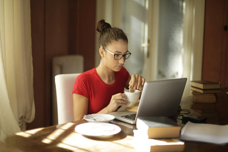 A young attractive female student reading eating cookies and looking at the laptop at home