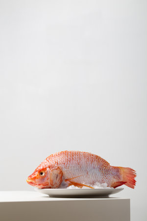 A vertical closeup of a raw red tilapia on a plate with ice on the table under the lights