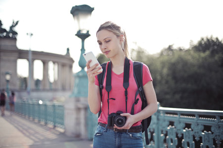 A shallow focus shot of a young female doing a city tour and holding in hands a smartphone and a cameraの写真素材