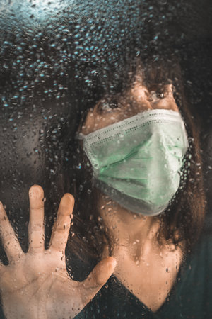 A woman with a medical mask looking out the window glass on a rainy day during the quarantineの写真素材