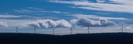 A panoramic shot of wind turbines on a mountain top against a cloudy blue sky with foreground trees...