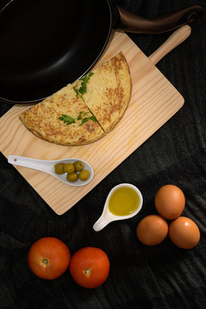 A vertical high angle closeup shot of two omelet pieces on a wooden cooking desk with olives, egg yolk, tomatoes, and fresh eggs on the sidesの写真素材