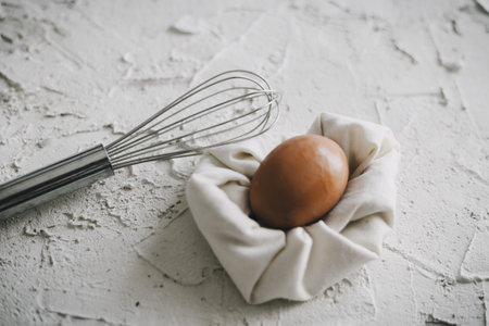 A hard-boiled egg on a white towel next to a silver whisk on a white surface