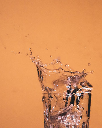 A vertical shot of water splashing out of a glass on an orange backgroundの写真素材