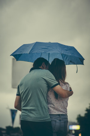 A vertical shot of a male hugging a female under a blue umbrella captured on a rainy day