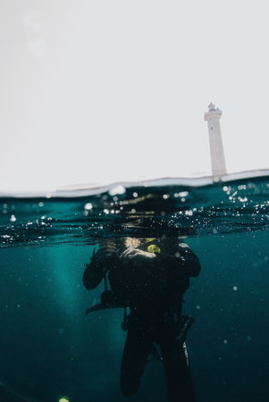 A vertical shot of a diver in the turquoise water and a lighthouse in the backgroundの写真素材