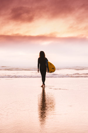 A vertical shot of a surfing woman standing on the hot sand and gleaming under the sunset