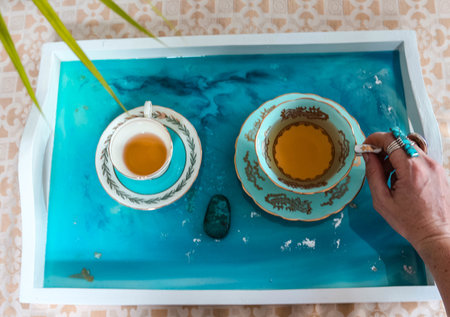 A high angle shot of a female hand grabbing a teacup from a tray with epoxy resin art with blue alcohol inksの写真素材