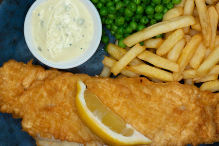 A closeup shot of french fries and fried fish with peas and sauce on a plate under the lights