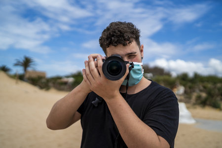 A male photographer wearing a facemask and taking a picture at the beach under the sunlight