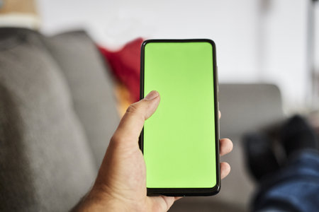 A closeup shot of a man holding a smartphone with a green screen whilst sitting on a gray sofa