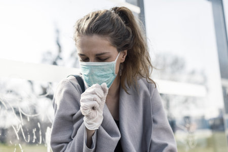 A young female wearing a facemask and coughing at a bus stop under the sunlight - COVID-19の写真素材