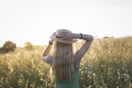 A beautiful shot of a blonde young female wearing a hat standing in the field at sunsetの写真素材
