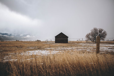 An old wooden barn at a field surrounded with a fence under a cloudy sky
