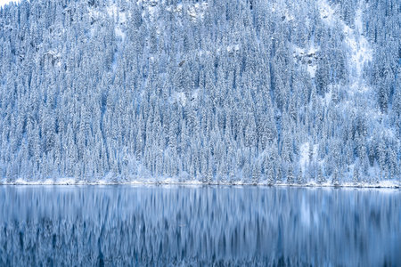 A beautiful shot of a calm lake with forested mountains covered in snowの写真素材