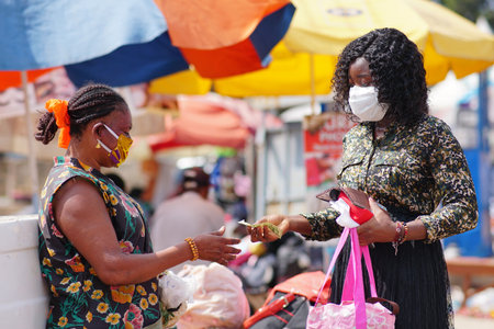 Young black woman paying money to a vendor for a purchased item in the market. Two women wearing locally made mask and surgical mask on the street.の写真素材