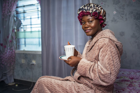 Beautiful Black girl wearing satin bonnet and morning coat, holding a cup and side plate. smiling and looking at camera - concept on Africa millennialの写真素材