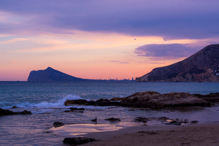The wavy ocean hitting the rocky beach surrounded by mountains under the pink cloudy skyの写真素材