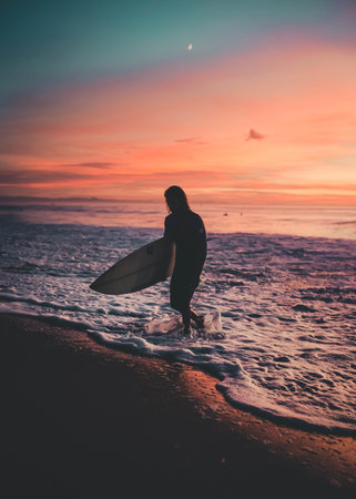 A surfer with a board walking out of the sea during sunsetの写真素材