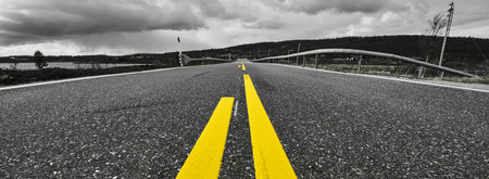 A grayscale shot of a road with yellow lines leading to mountains