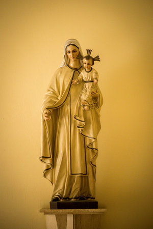 A vertical shot of a statue of Mary, mother of Jesus, holding Jesus Christ with a yellow wall behind
