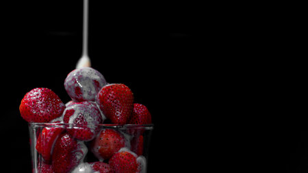 A closeup shot of fresh strawberries in a glass bowl with milk pouring on a black backgroundの写真素材