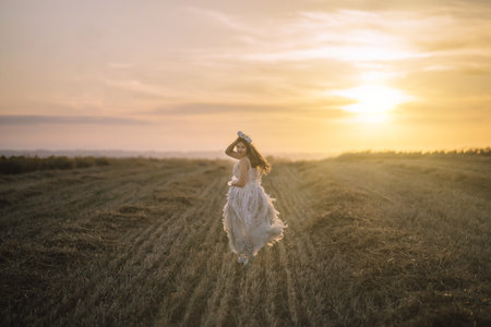 A young brunette female in a white dress looking back and holding a flower crown to her head while running towards the sunset in a fieldの写真素材