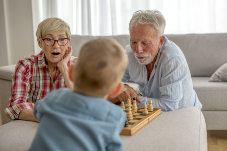 A selective focus shot of grandmother with short blond hair and red glasses and grandfather with gray hair and beard playing chess with their grandsonの写真素材