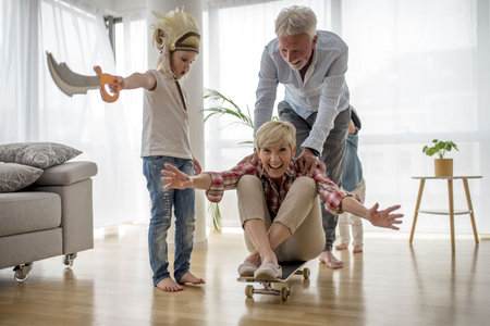 A caucasian grandfather pushing a caucasian grandmother sitting on a skateboard in their home while grandson wearing pirate costume looksの写真素材