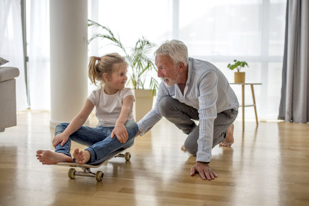 A caucasian blonde female child wearing jeans and a t-shirt sitting on a skateboard looking at her grandfather pushing her inside a houseの写真素材